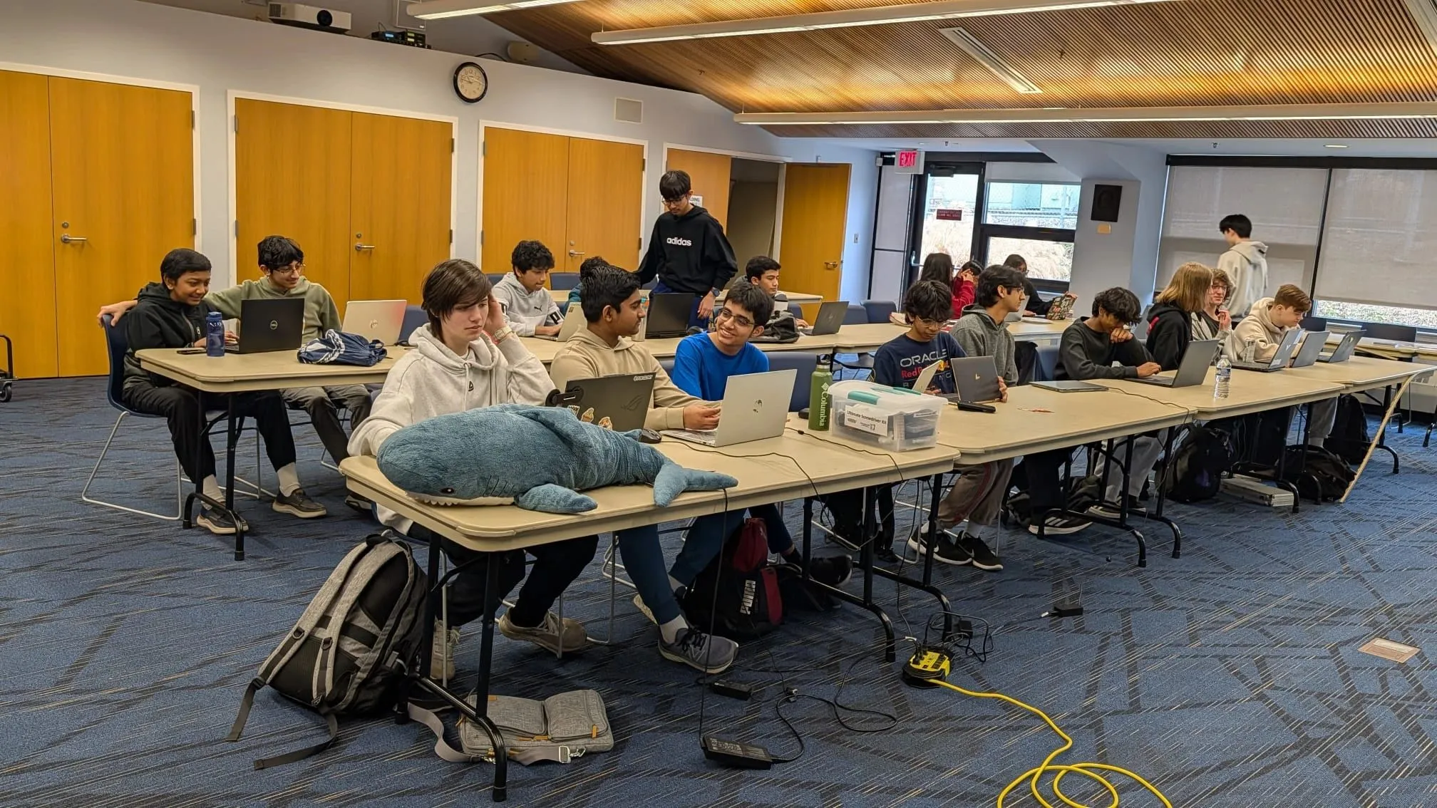 Hackathon workroom: rows of students heads-down over laptops, one desk guarded by a plush shark.